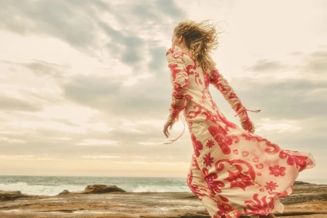 Woman in a long patterned cream and red dress walking along a rocky coast under a cloudy sky.