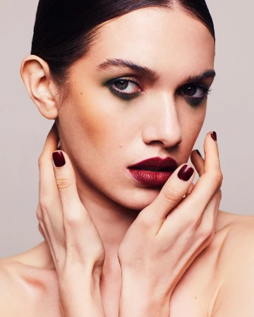 Close-up portrait of a woman with dark hair, bold plum lipstick, green eyeshadow, and matching dark nails, holding her hands near her face.