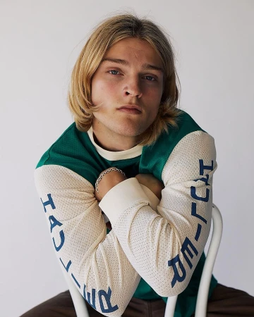 Young man with long blonde hair wearing a green and white long-sleeve top seated on a white chair.