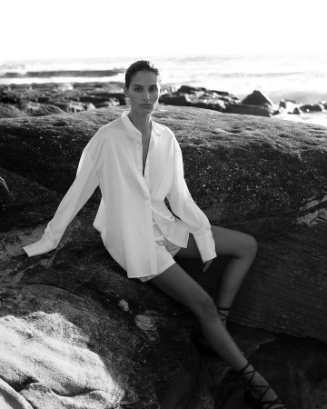 Model wearing an oversized white button-down shirt sitting on dark coastal rocks by the ocean in a black and white portrait.