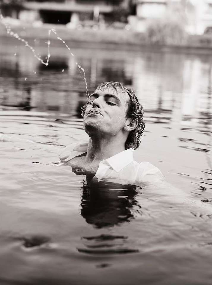 Monochrome close-up of a man in water, wearing a white shirt, with water spraying from his mouth towards the sky in a monochrome outdoor setting.
