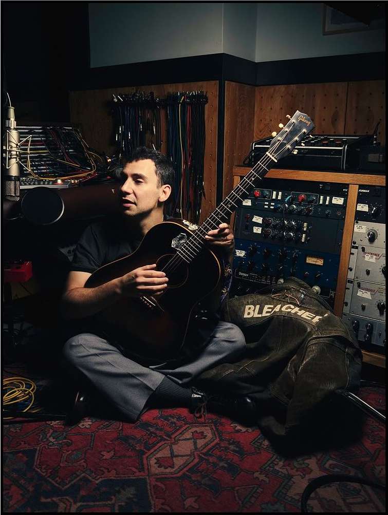 Man sitting cross-legged in a music studio, holding a guitar and singing into a microphone, surrounded by sound equipment.
