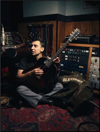 Man sitting cross-legged in a music studio, holding a guitar and singing into a microphone, surrounded by sound equipment.