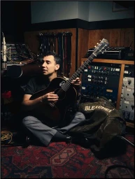 Musician seated cross-legged on a patterned rug holding an acoustic guitar inside a recording studio