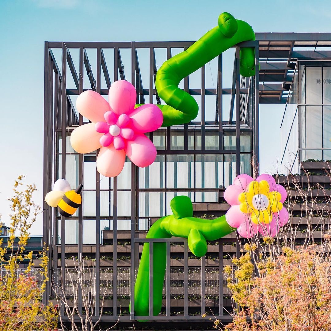 Inflatable flowers and a bee adorn an urban structure against a blue sky, creating a whimsical, playful outdoor installation.