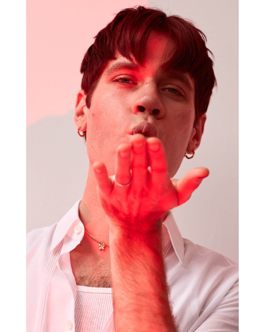 Portrait of a young adult man with short brown hair wearing a white shirt, blowing a kiss in a studio with red lighting.