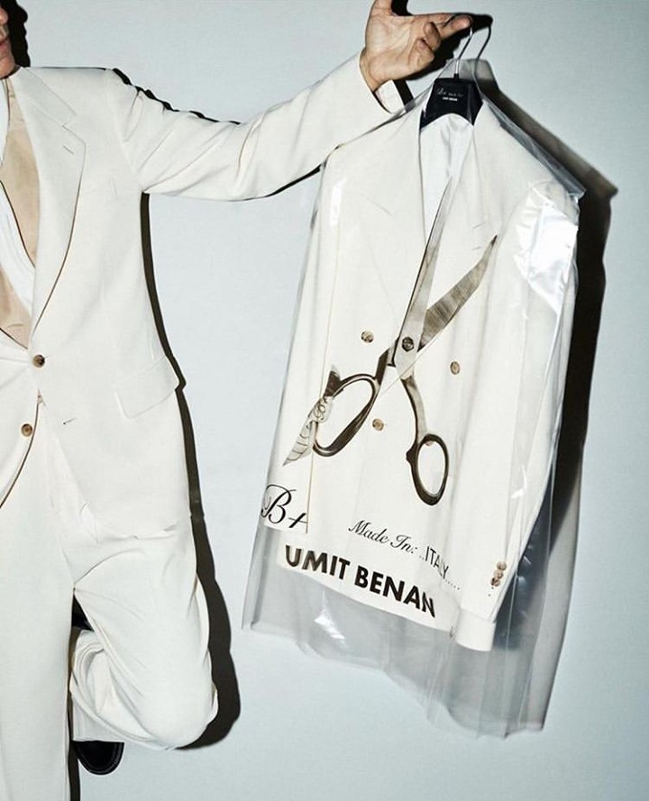 Person in a white suit holding a suit in a garment bag featuring a scissors design, against a plain backdrop, showcasing fashion and style.