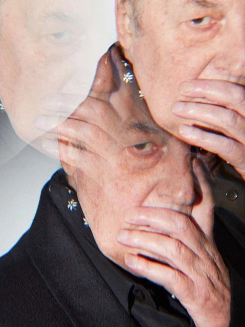 Close-up portrait of an elderly adult male with fair skin, wearing a black button-down shirt and patterned scarf, covering his mouth with his hand.
