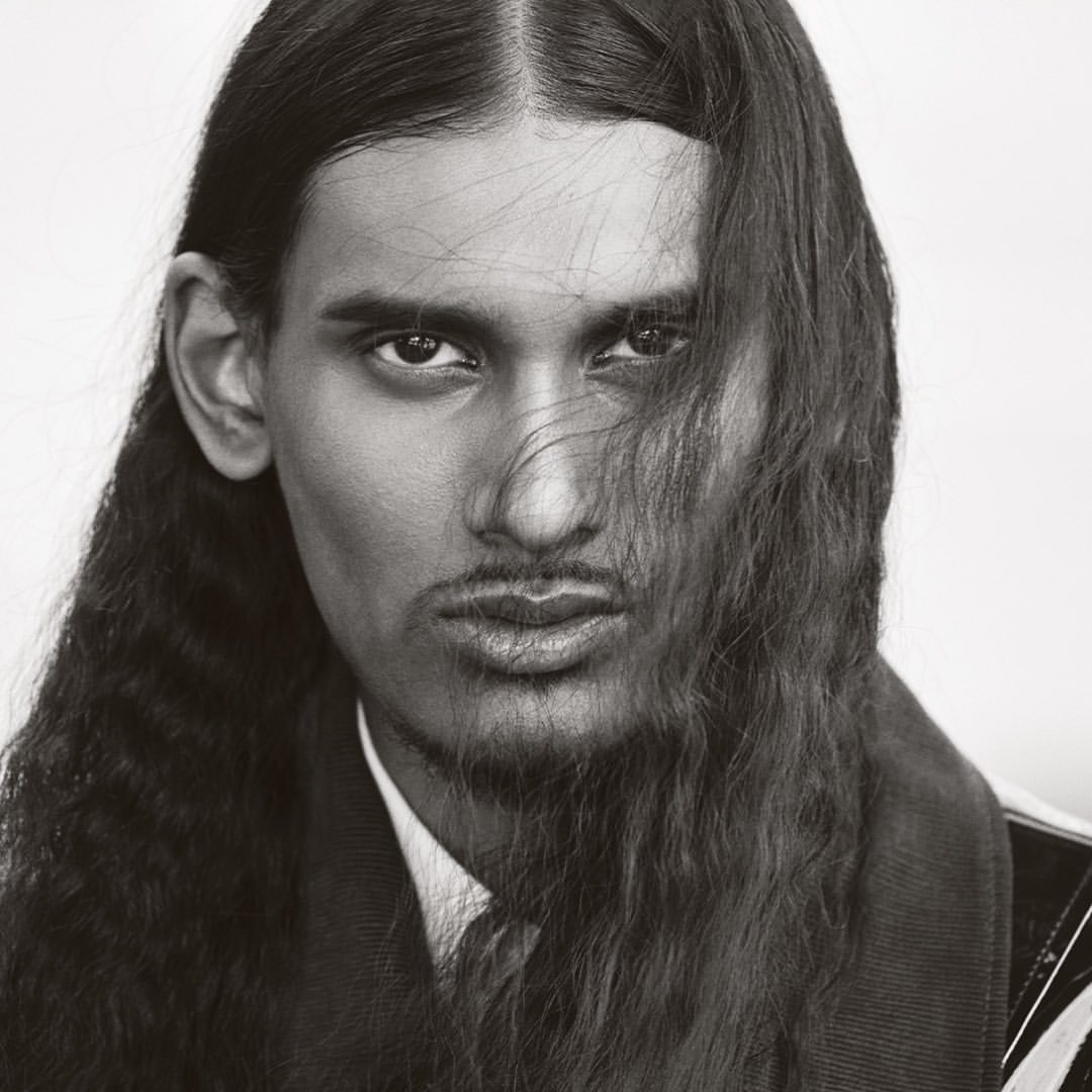 Close-up portrait of a man with long dark hair and facial hair, wearing a collared shirt and jacket against a plain background.