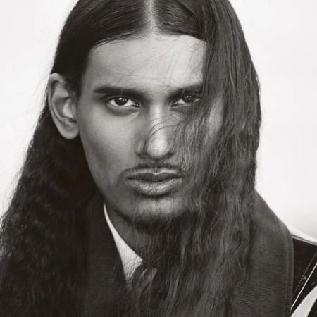 Close-up portrait of a man with long dark hair and facial hair, wearing a collared shirt and jacket against a plain background.