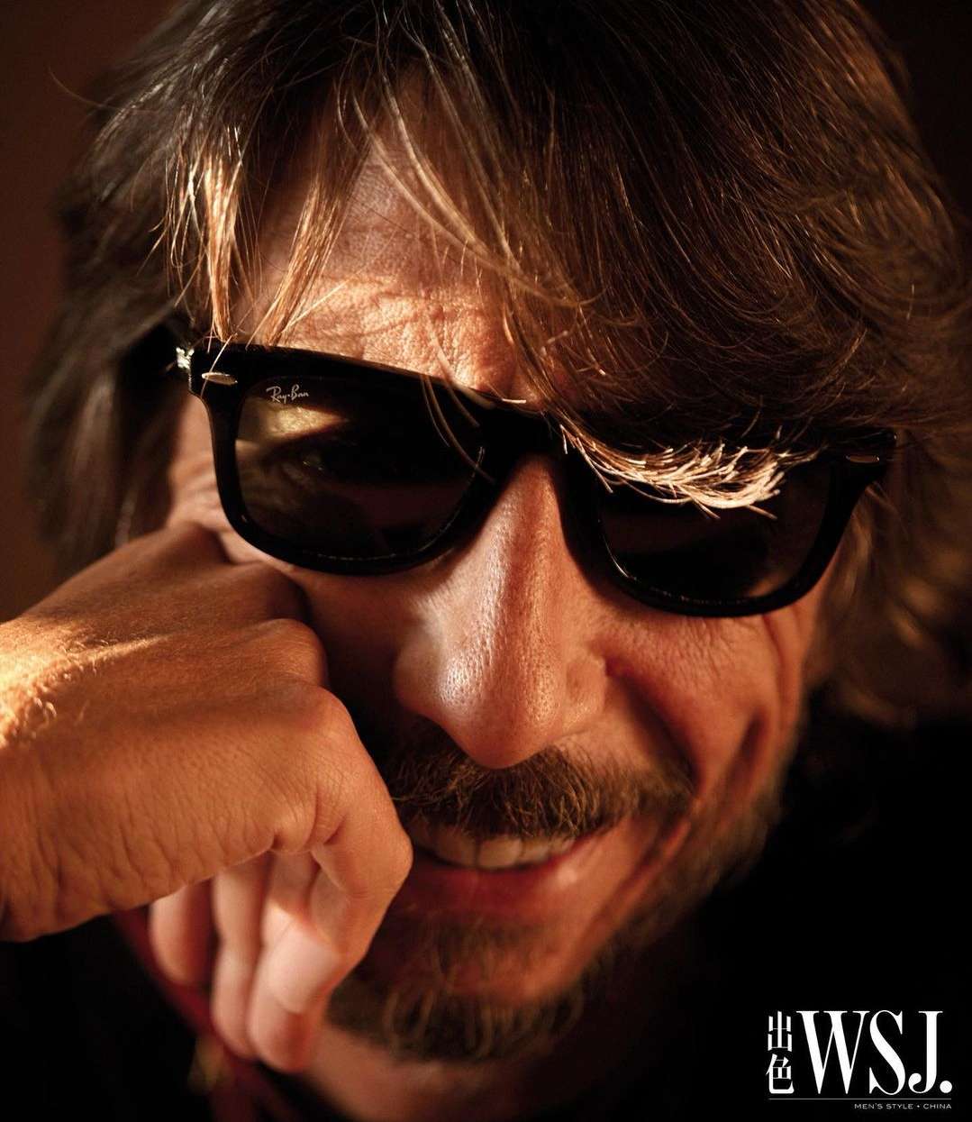 Close-up portrait of a man with medium brown hair wearing black sunglasses, resting his chin on his hand with a slight smile.