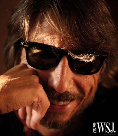 Close-up portrait of a man with medium brown hair wearing black sunglasses, resting his chin on his hand with a slight smile.