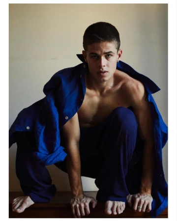 Young man with short brown hair crouching barefoot wearing loose blue trousers and an unbuttoned blue shirt
