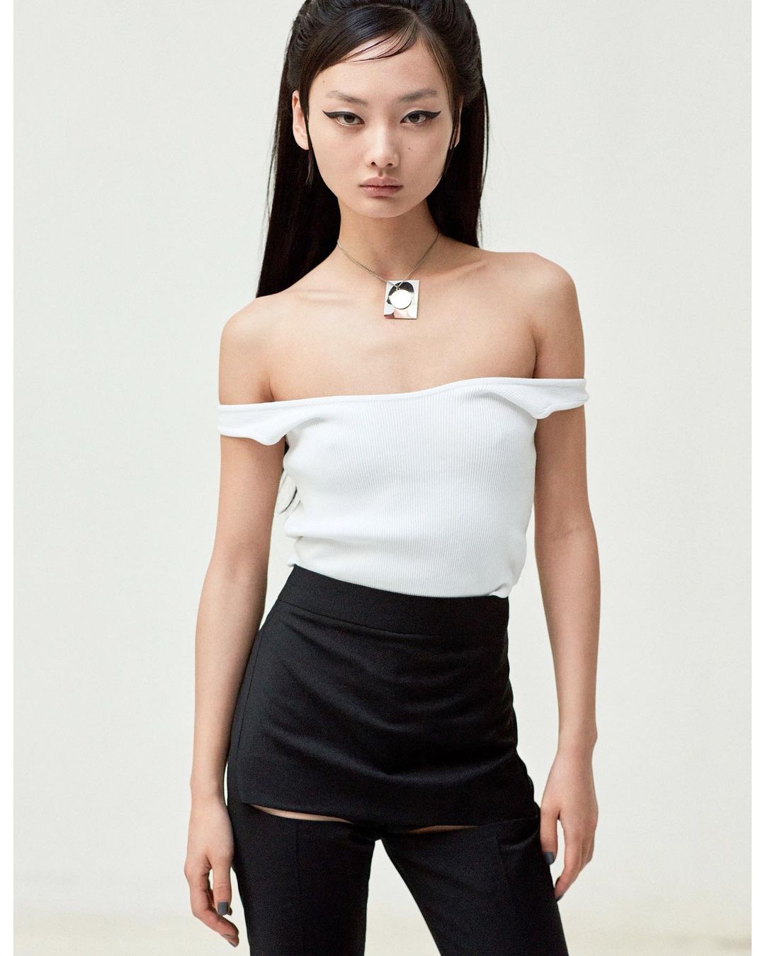 Studio shot of an adult woman wearing a white, off-the-shoulder top, black bottoms, and a silver necklace against a neutral background.