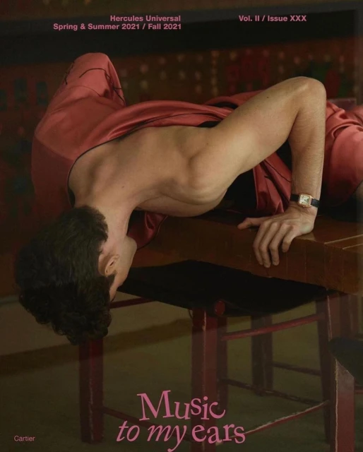 Man in red loungewear leans on a wooden table in a studio with dark colors and soft lighting, showcasing a wristwatch and stylized pose.