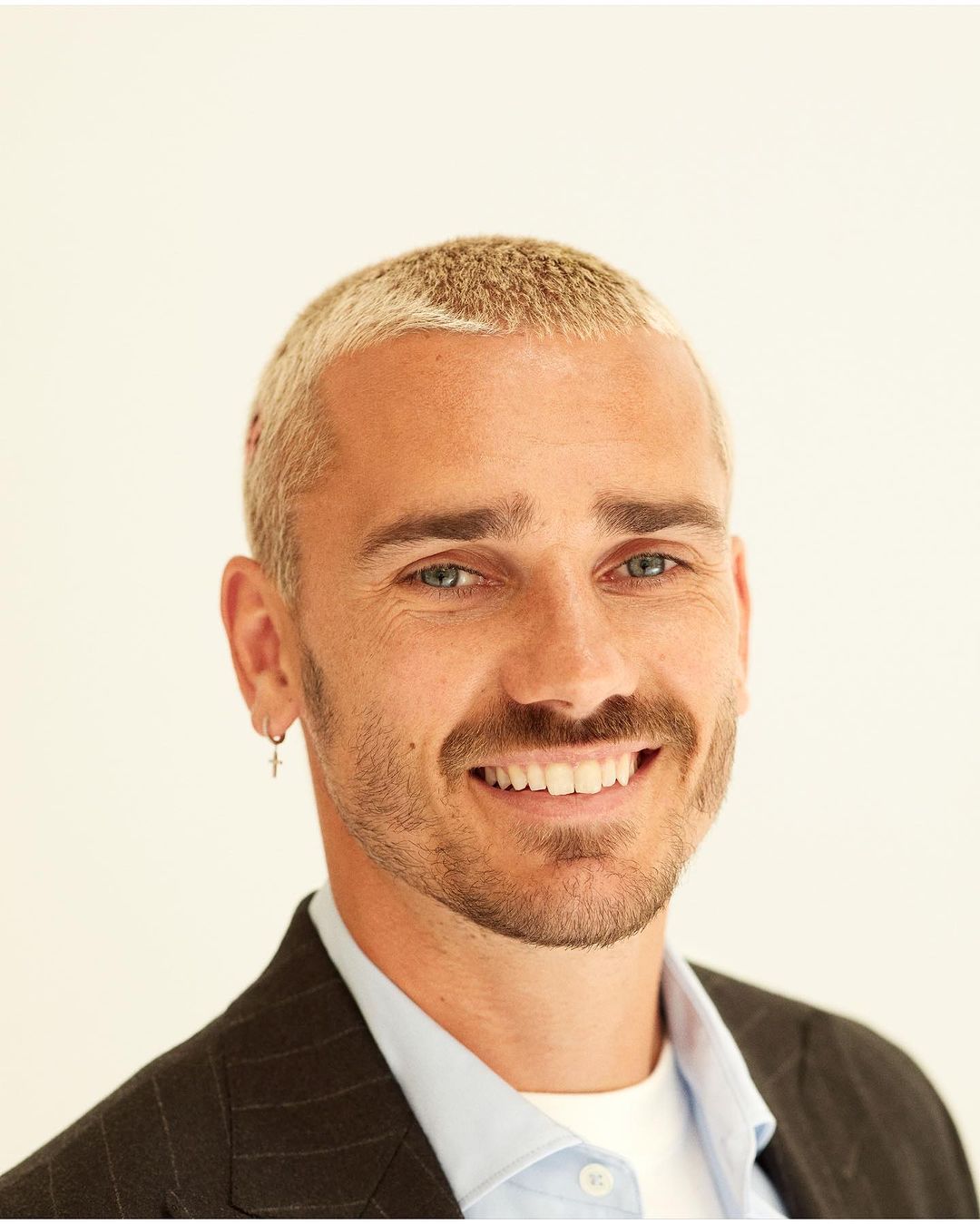 Close-up portrait of a smiling adult male with bleached blond hair, wearing a blazer and light blue shirt against a white background.