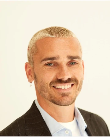 Close-up portrait of a smiling adult male with bleached blond hair, wearing a blazer and light blue shirt against a white background.