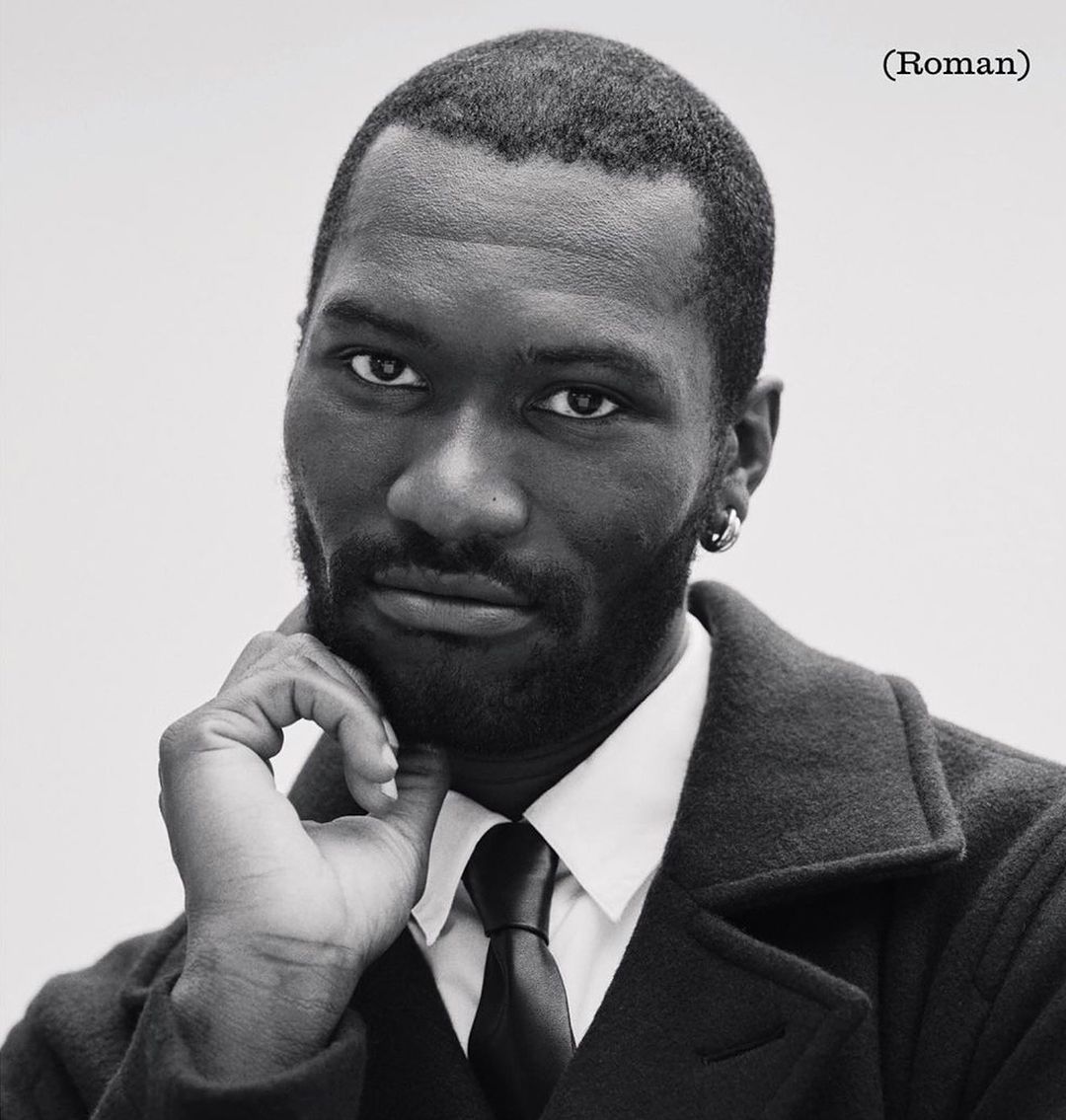 Black and white portrait of a Black man wearing a dark coat, white shirt, and tie, posed with hand to chin against a plain background.