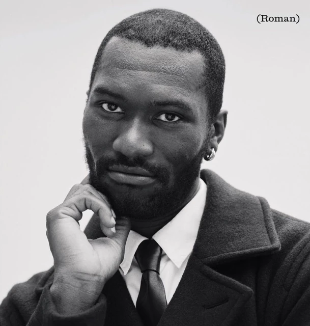 Black and white portrait of a Black man wearing a dark coat, white shirt, and tie, posed with hand to chin against a plain background.