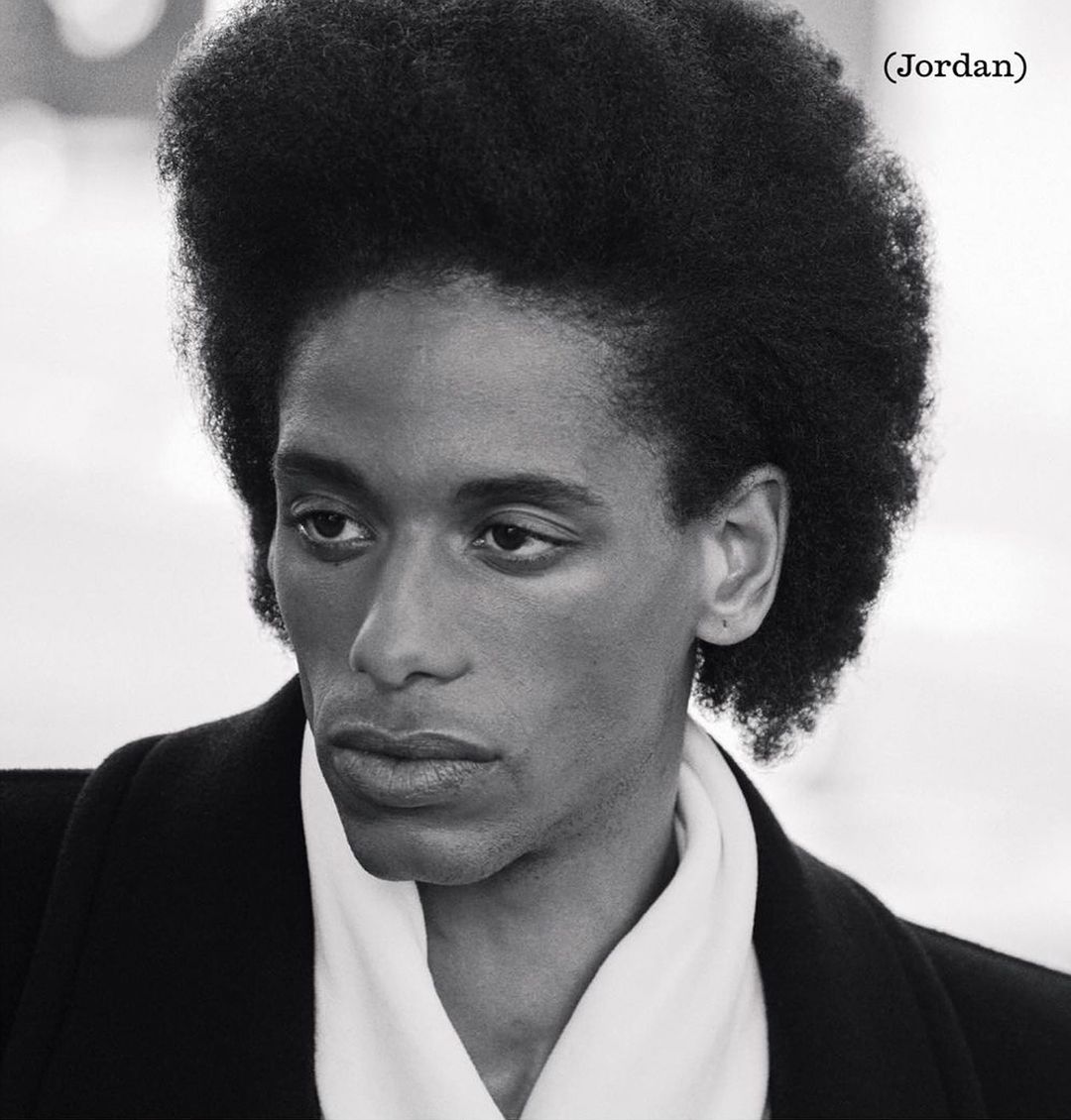 Striking black and white portrait of a young adult with distinctive afro hairstyle wearing an elegant black coat and white scarf.
