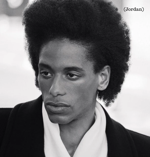Striking black and white portrait of a young adult with distinctive afro hairstyle wearing an elegant black coat and white scarf.