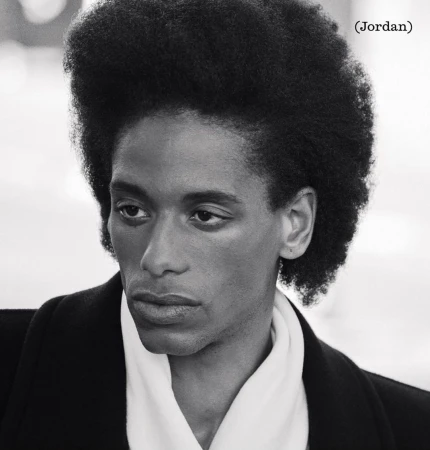 Striking black and white portrait of a young adult with distinctive afro hairstyle wearing an elegant black coat and white scarf.