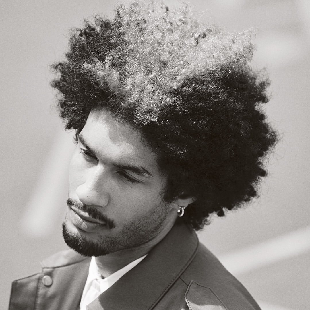 Close-up portrait of a man with textured dark hair and a mustache wearing a collared jacket and hoop earring.