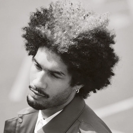 Close-up portrait of a man with textured dark hair and a mustache wearing a collared jacket and hoop earring.