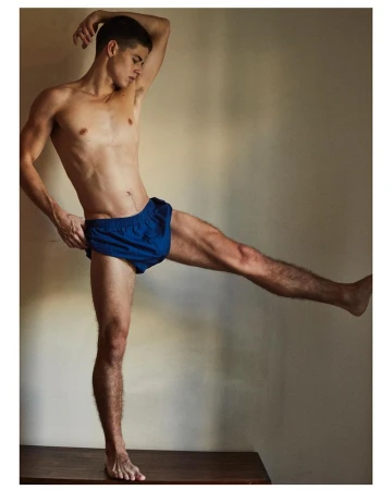 Shirtless male model wearing blue athletic shorts posing with one leg raised, photographed by Daniel Riera in Paris.