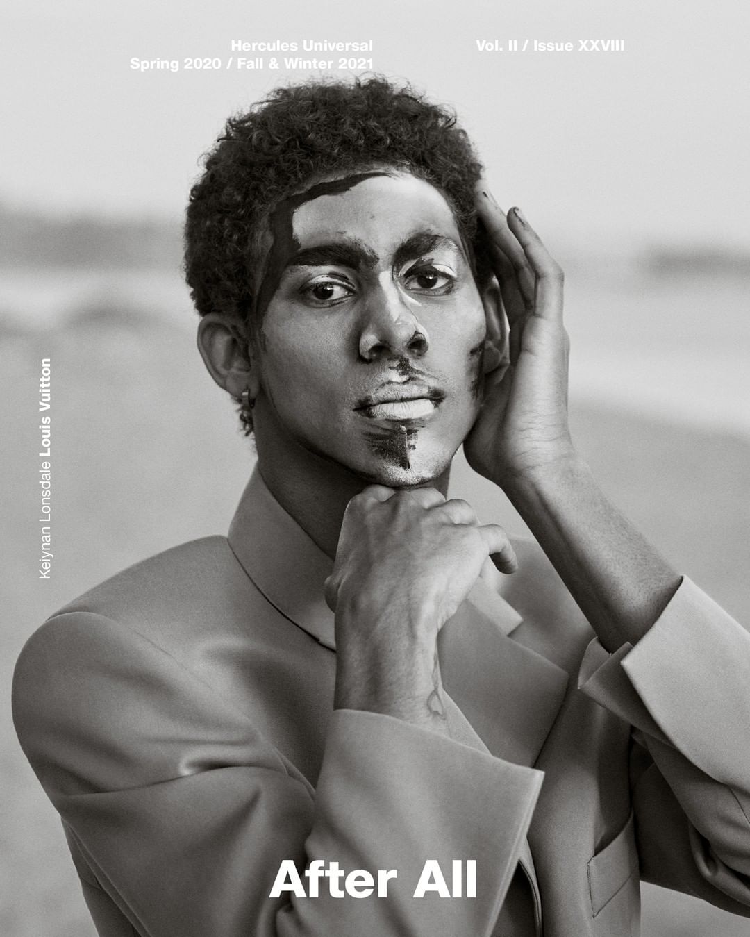 Portrait of a biracial male model with black and white paint on his face and hands in a tan suit jacket, against a neutral outdoor backdrop.