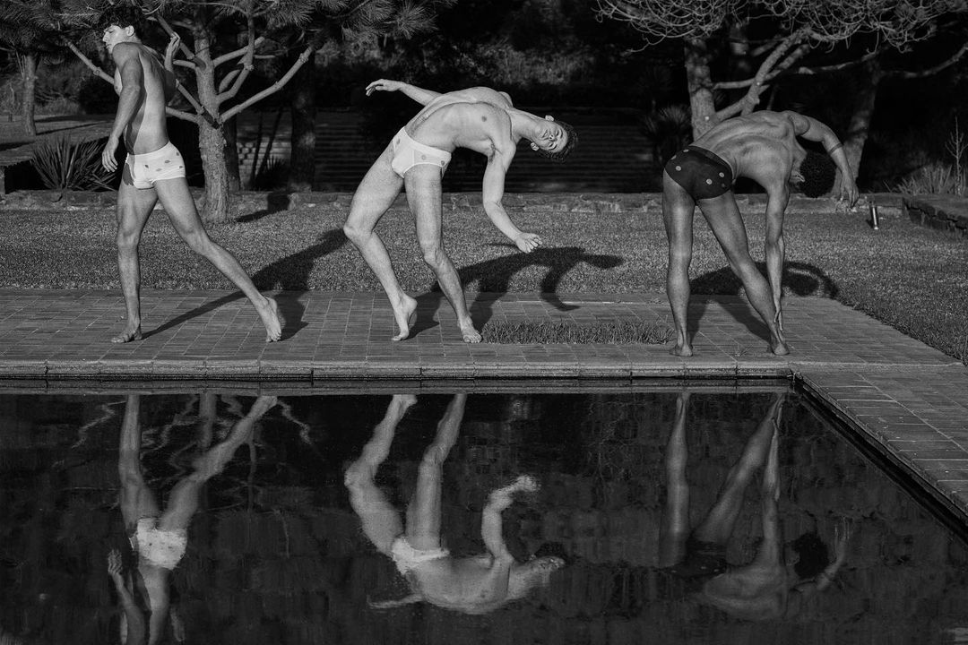 Monochrome image of three male models posing beside a swimming pool, with sharp reflections visible in the dark water.