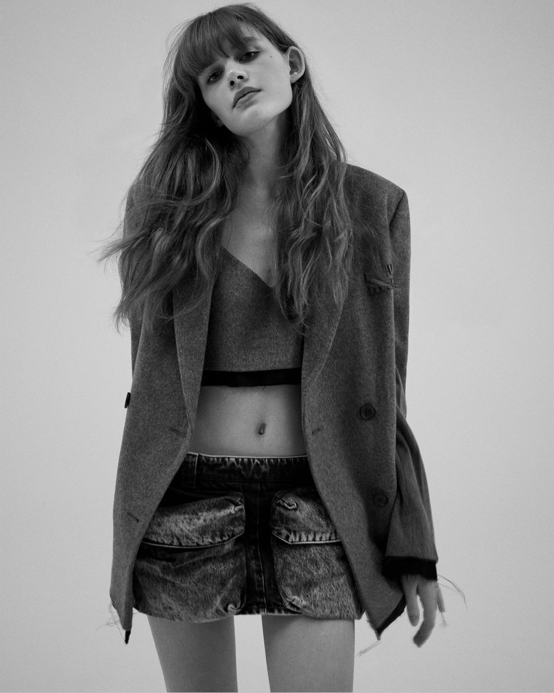 A young woman in a gray blazer, crop top, and denim mini skirt poses against a light backdrop in a studio shot, medium angle.