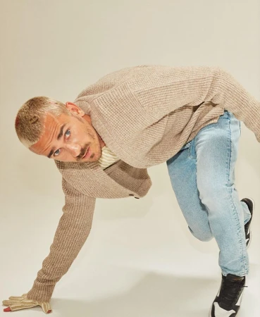 Indoor shot of a blonde adult male in a beige cardigan and light blue jeans, leaning forward with one hand on the floor.