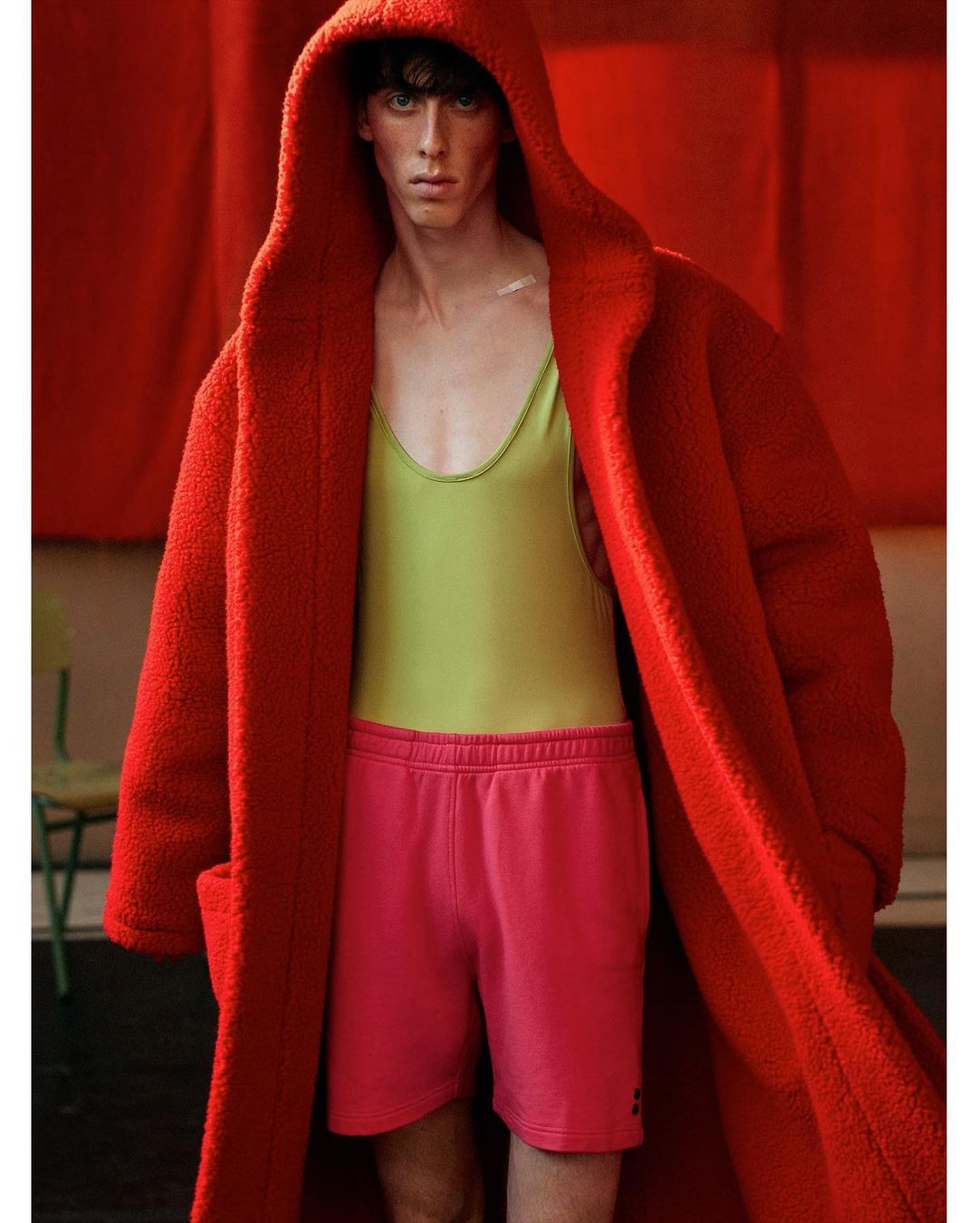 Fashion shot of adult male in a red hooded coat and pink shorts standing indoors with red backdrop and chair. Model is posing.