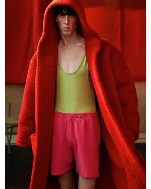 Fashion shot of adult male in a red hooded coat and pink shorts standing indoors with red backdrop and chair. Model is posing.