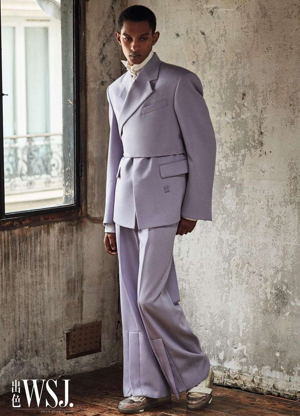 Male fashion model in a lilac suit with wide-legged pants poses near a window with a distressed wall in the background.