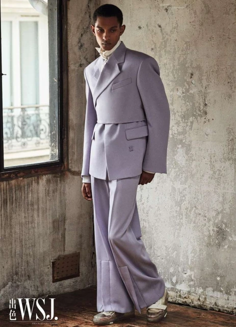 Male fashion model in a lilac suit with wide-legged pants poses near a window with a distressed wall in the background.