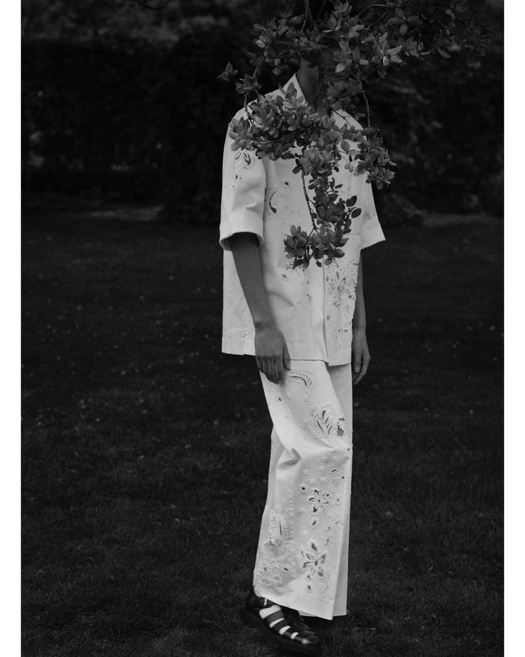 Monochromatic fashion shot of an adult wearing an eyelet-design outfit with leaves obscuring the face in a grassy outdoor setting.