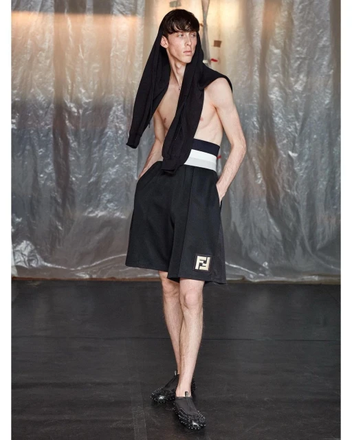 A slender man in black shorts and textured shoes stands in a hooded top indoors against a shimmering plastic backdrop, hands in pockets.