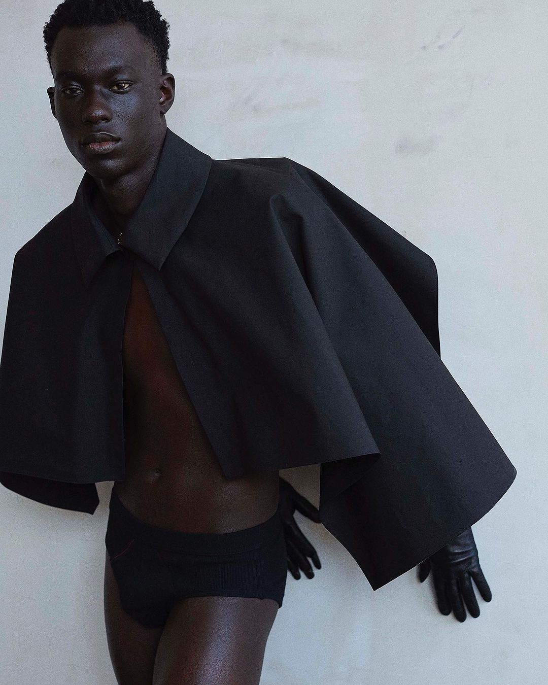 A dark-skinned male model poses indoors, wearing a black top and briefs against a textured wall, showcasing fashion and editorial style.