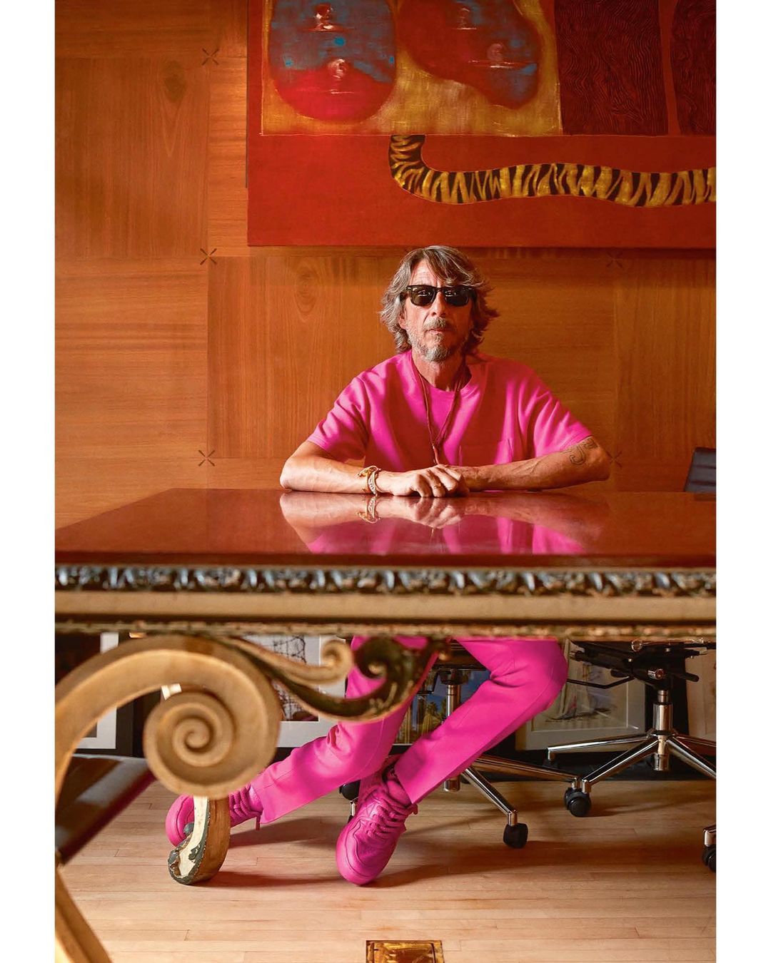Adult man in pink outfit sits at ornate wood desk in office setting with abstract painting in background, wearing sunglasses.