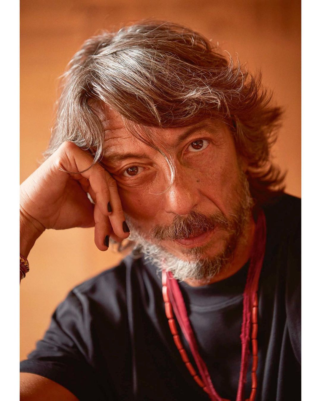 Close-up studio shot of a gray-haired adult male wearing a black t-shirt and red beaded necklaces, with a pensive facial expression.