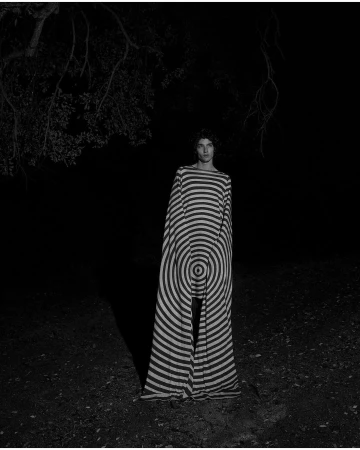 Monochrome portrait of an adult woman in a striped dress standing outdoors at night, framed by trees. Fashion or editorial photography.