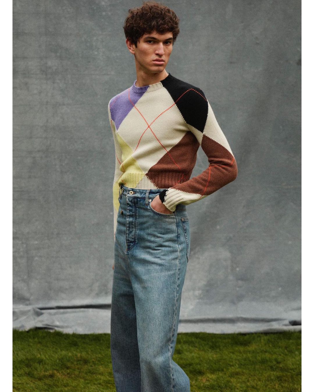 A young adult male fashion model in a patterned sweater and blue jeans stands in a photo studio against a gray backdrop with greenery.
