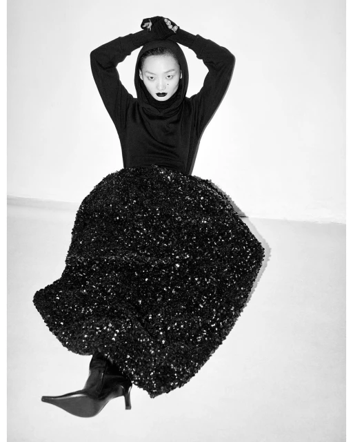 Elegant portrait of an Asian woman wearing a black hooded top and a sequined skirt, posing against a neutral background in a studio.