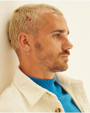 Headshot of soccer player Antoine Griezmann, side profile, wearing a blue turtleneck and cream-colored corduroy jacket, neutral background.
