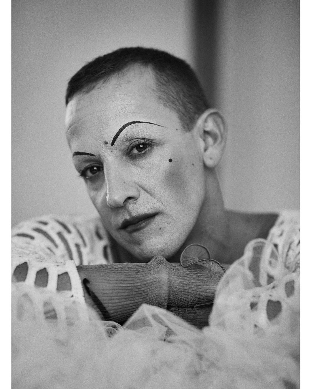 Monochrome portrait of a person with theatrical makeup leaning on a textured prop, wearing a lace-like white top against a plain background.