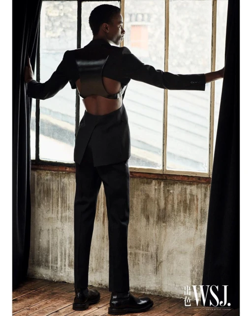 Androgynous fashion model posing in a black suit with harness, standing near a window with dramatic lighting and textured background.