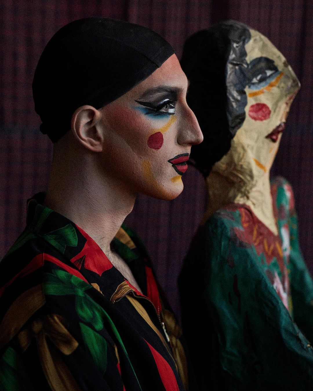Portrait of a person with stage makeup and a black head covering next to a stylized mannequin against a dark, textured background.