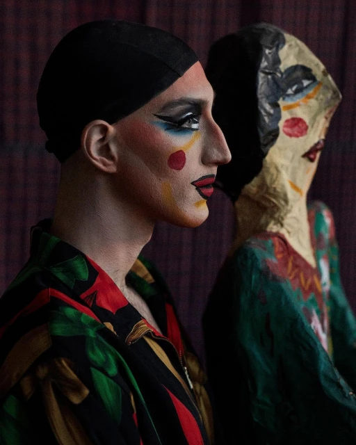 Portrait of a person with stage makeup and a black head covering next to a stylized mannequin against a dark, textured background.